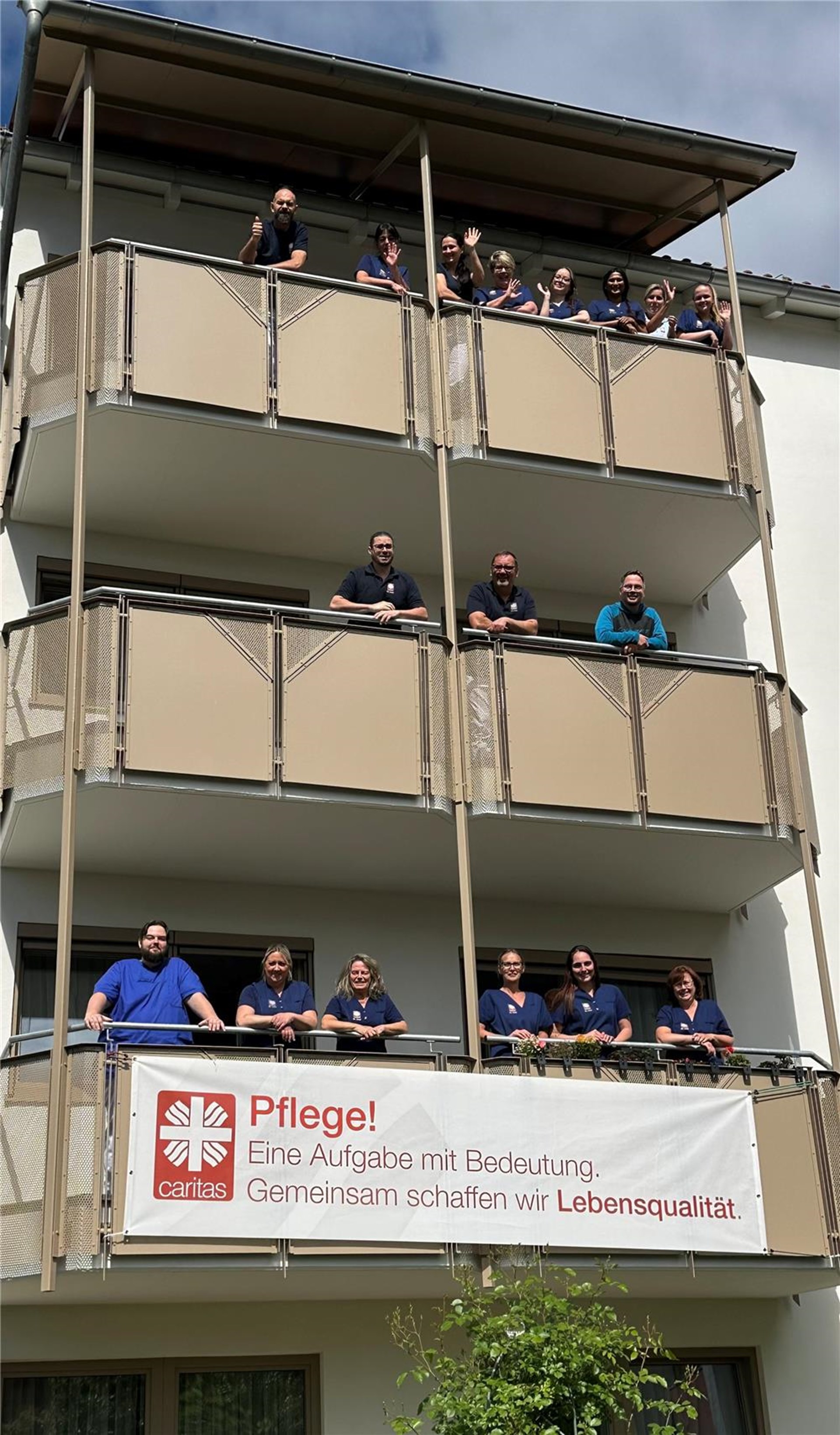 Pflegeteam steht und winkt von mehreren Balkonen eines Pflegeheims zum Tag der Pflegenden. Auf dem Banner steht „Pflege. Eine Aufgabe mit Bedeutung. Gemeinsam schaffen wir Lebensqualität.“