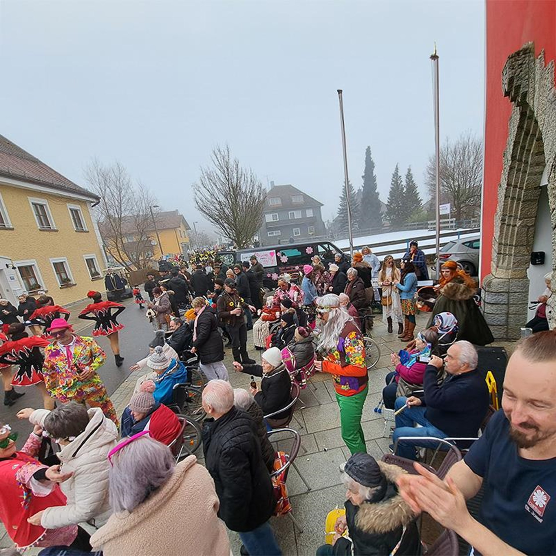 auf dem Bild ist eine Strasse zu sehen, am Strassenrand stehen viele Menschen, auch mit Rollatoren und Rollstühlen und beaobachten den Faschingszug der auf der Strasse vorbeizieht